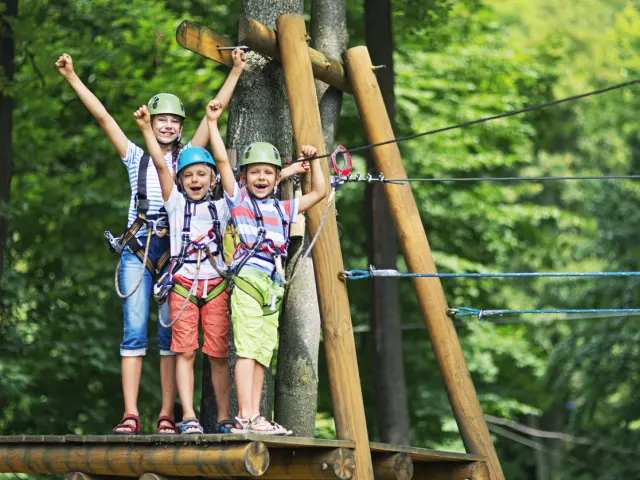 Meisje van 10 met haar broertjes van 7, met helmen op, op een houten platform met een tokkelbaan in het avonturenpark. De kinderen lachen naar de camera en applaudisseren.