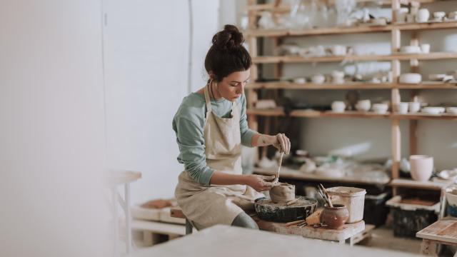 Mooie jonge vrouw met pottenbakkersinstrument om te schrapen, glad te maken, vorm te geven en te beeldhouwen. Dame zittend op bank met pottenbakkersschijf en kleipot aan het maken