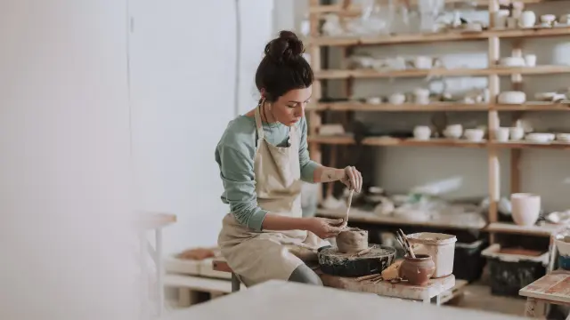 Beautiful young woman holding pottery instrument for scraping, smoothing, shaping and sculpting. Lady siting on bench with pottery wheel and making clay pot