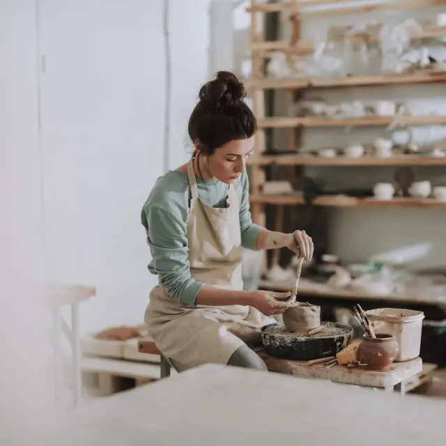 Mooie jonge vrouw met pottenbakkersinstrument om te schrapen, glad te maken, vorm te geven en te beeldhouwen. Dame zittend op bank met pottenbakkersschijf en kleipot aan het maken