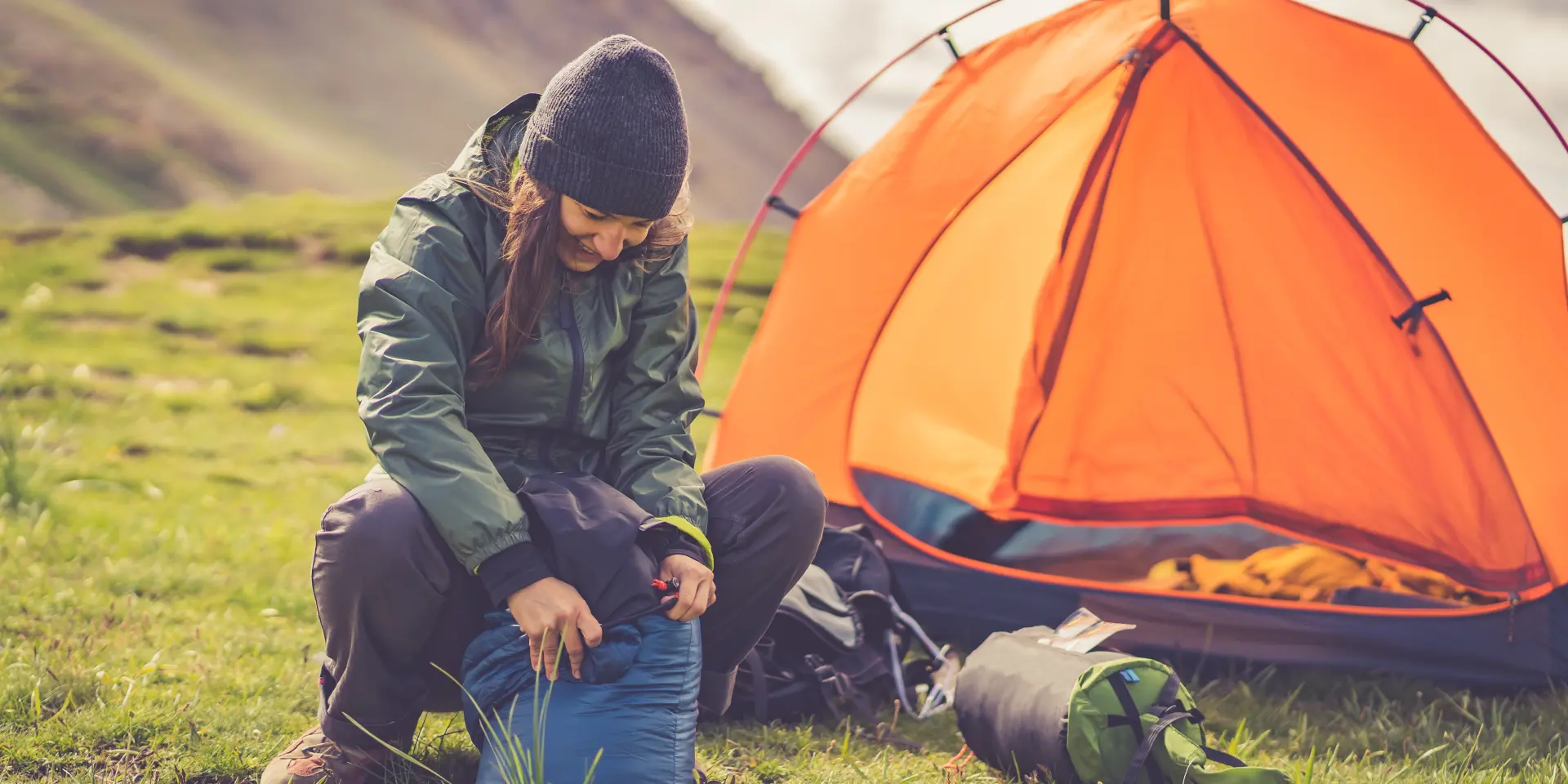 alpine woman folding sleeping bag and other accessories for camping. In the background orange tent and equipment for outdoor activities.